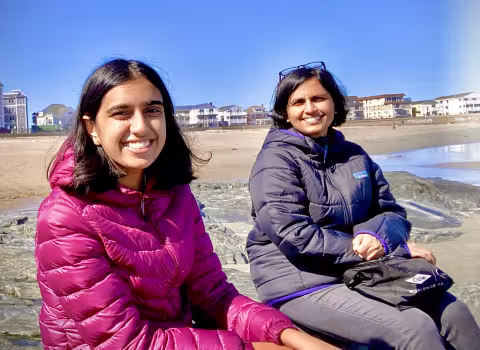 Two smiling researchers wearing jackets sit on rocks at a sunny beach with shoreline homes and the ocean in the background.